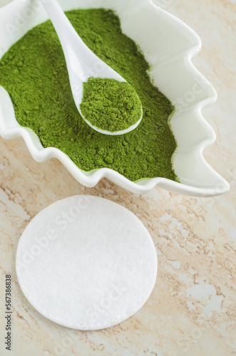 White porcelain bowl with matcha green tea powder (henna, seaweed) and cotton pad. Natural beauty treatment and spa. Homemade matcha mask recipe. Selective focus.