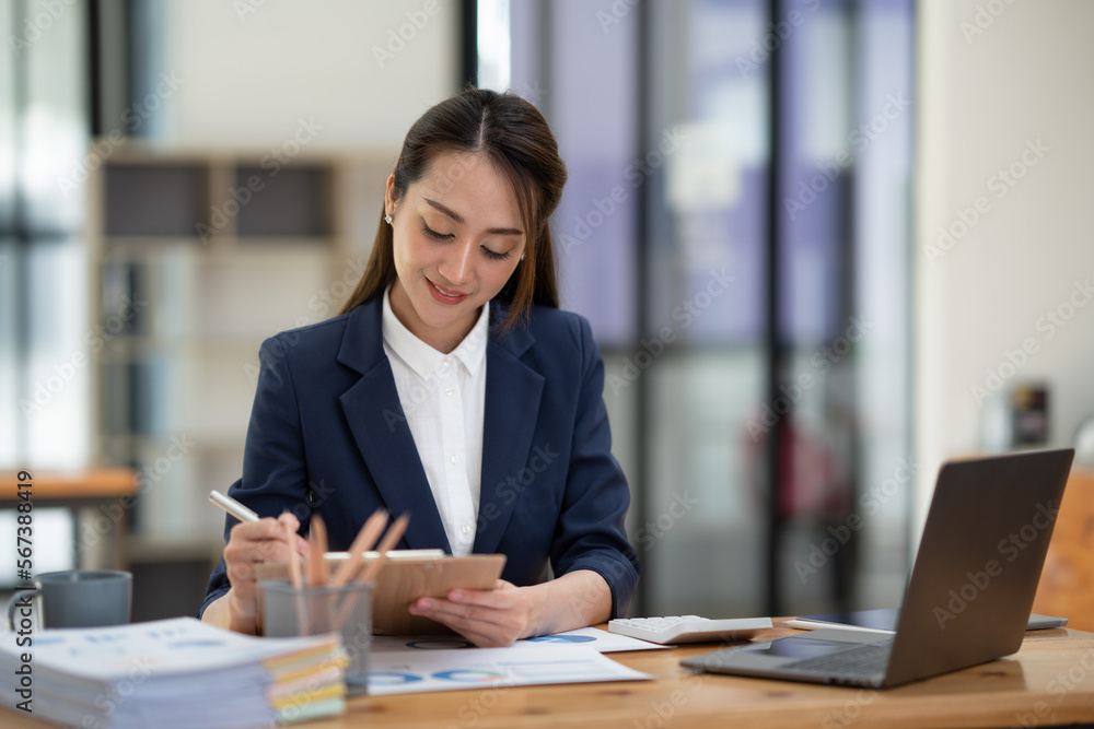 Beautiful attractive Asian businesswoman holding a clipboard of documents sitting analytically analyzing various marketing management information the finances were at her desk in the office at ease. Beautiful attractive Asian businesswoman holding a clipboard of documents sitting analytically analyzing various marketing management information the finances were at her desk in the office at ease.
