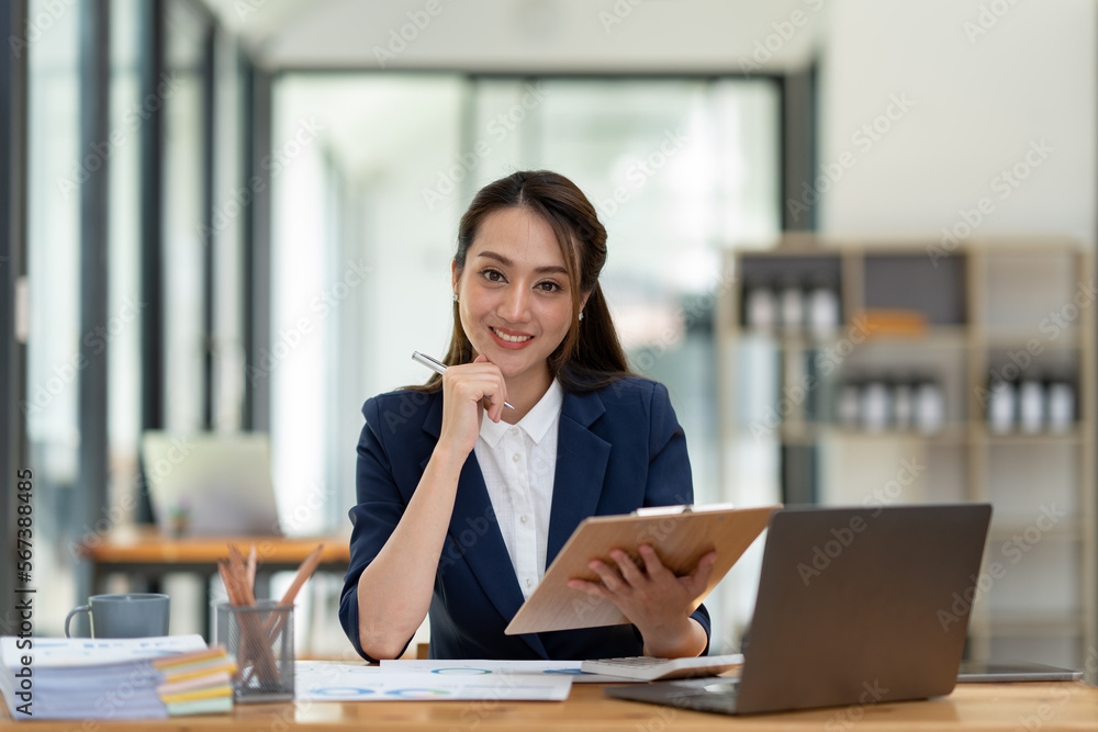 Beautiful attractive Asian businesswoman holding a clipboard of documents sitting analytically analyzing various marketing management information the finances were at her desk in the office at ease. Beautiful attractive Asian businesswoman holding a clipboard of documents sitting analytically analyzing various marketing management information the finances were at her desk in the office at ease.