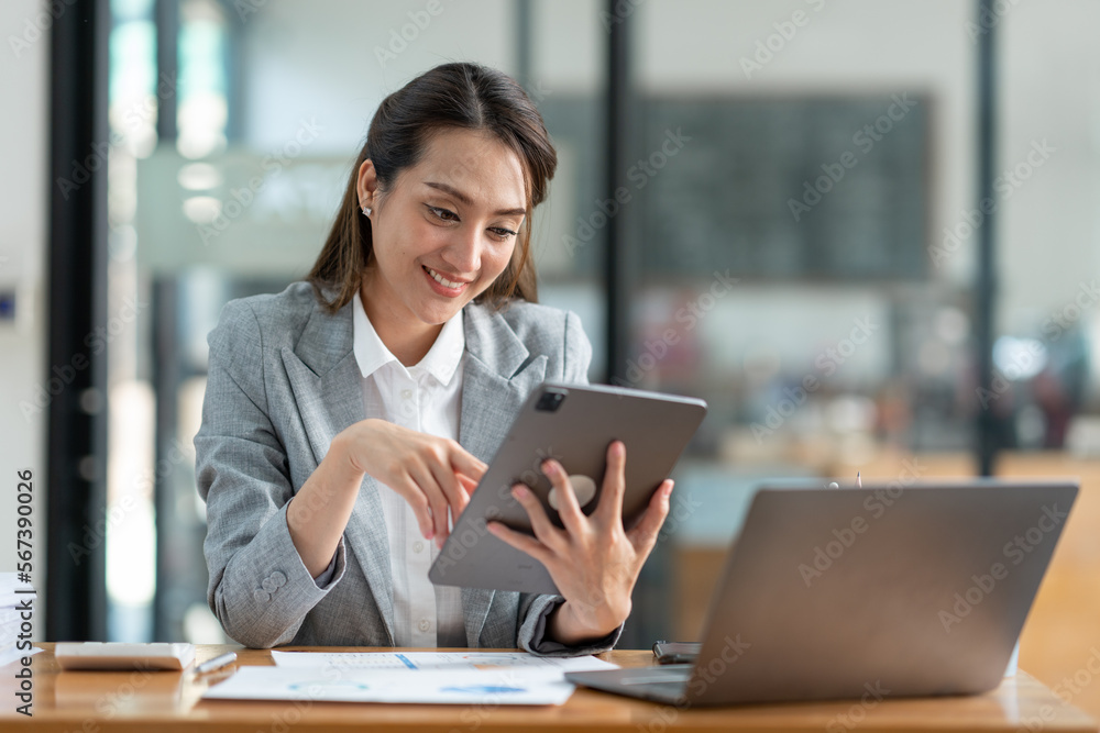 Asian businesswoman in a suit sitting holding iPad or tablet in contact See details and discuss business information happily in the office. Asian businesswoman in a suit sitting holding iPad or tablet in contact See details and discuss business information happily in the office.