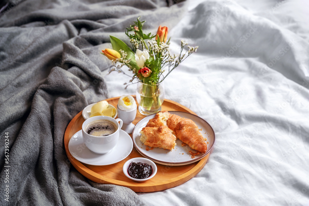 Wooden breakfast tray with croissant, coffee, jam, egg and a fresh flower bouquet served on bed with light linen and gray blanket, holiday or weekend morning, copy space - obrazy, fototapety, plakaty 