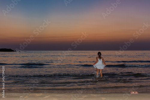 Little girl on the beach, wearing a dress and going into the sea during a summer sunrise