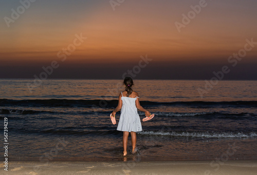 Girl from behind, in a dress, carrying flip flops while the sea wets her feet during a summer sunrise
