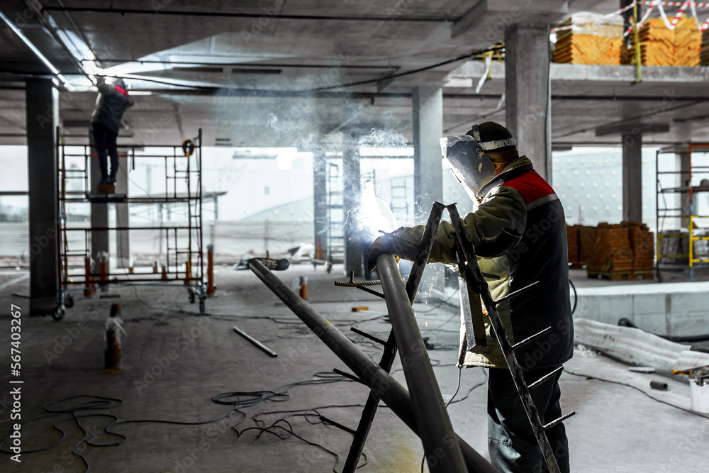 Construction of a production facility. Two handymen perform welding and grinding at their workplace in the workshop. Man in protective helmet and gear Construction of a production facility. Two handymen perform welding and grinding at their workplace in the workshop. Man in protective helmet and gear