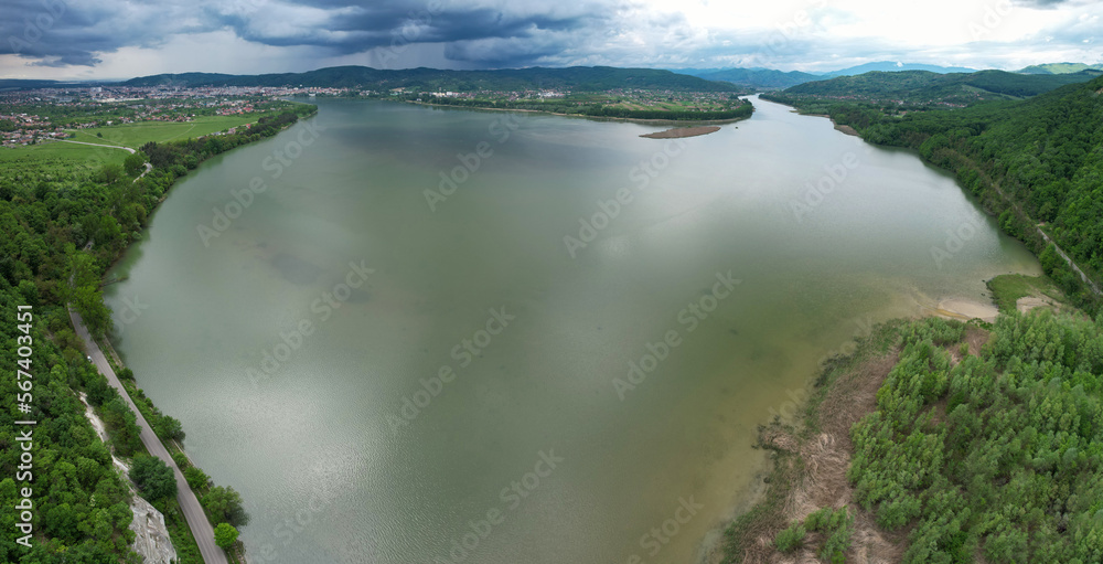 Naklejka premium Aerial drone panorama above a lake in Valcea County, Romania. Stormy weather, rain clouds are gathering above the surrounding forests. Several small villages can be seen on the lake's shores.