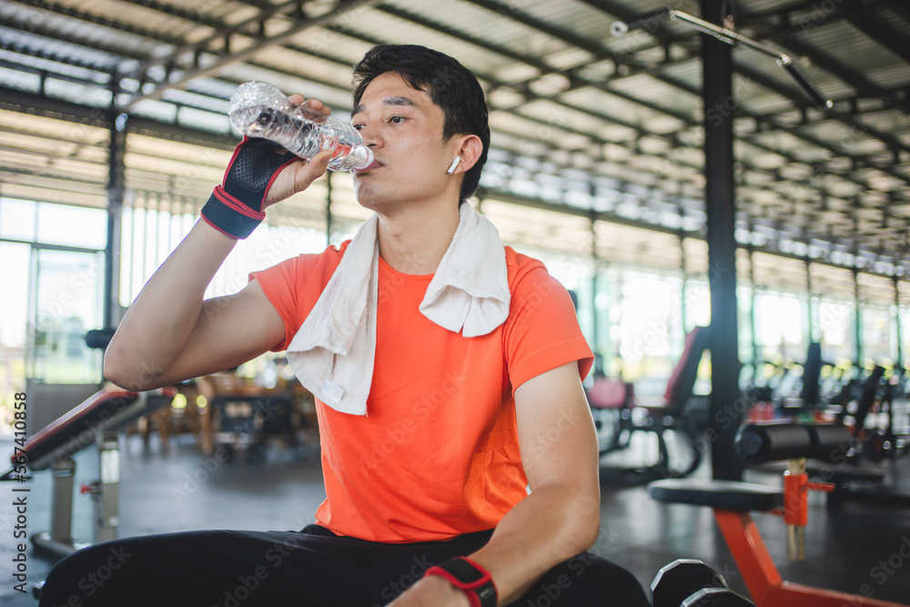Asian man is sitting on bench and drinking water to relax after workout in gym. Asian man is sitting on bench and drinking water to relax after workout in gym.