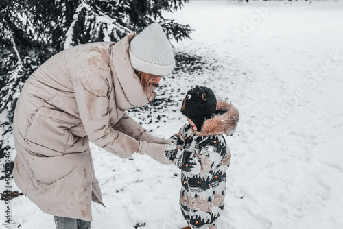 A girl in a beige coat teaches a child of 2 years to make snowballs