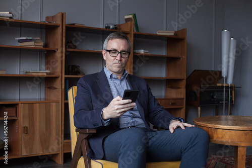 Confident elderly businessman in blue suit and glasses working and looking on his mobile phone indoor on office wall with boockshelf