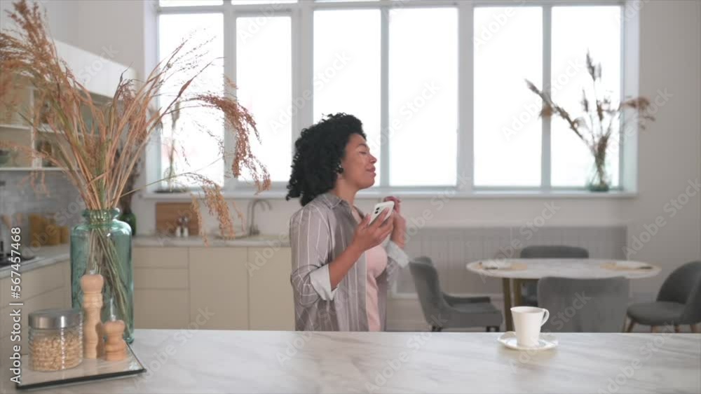 Portrait of a happy woman singing using phone as a microphone in the home kitchen, front view, slow motion. A young girl is dancing cheerfully and her curly hair is flying. Portrait of a happy woman singing using phone as a microphone in the home kitchen, front view, slow motion. A young girl is dancing cheerfully and her curly hair is flying.