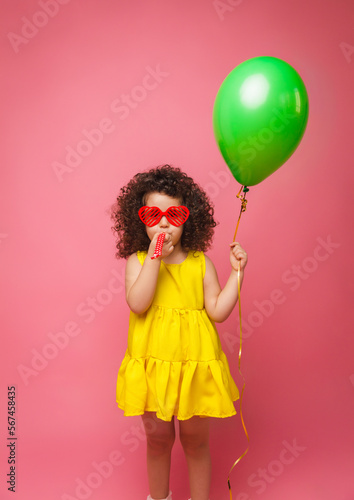 Happy birthday celebration with flying balloons of a charming cute little girl in a yellow dress isolated on a pink background.