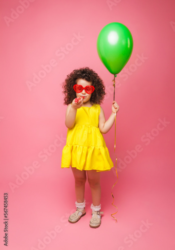 Happy birthday celebration with flying balloons of a charming cute little girl in a yellow dress isolated on a pink background.