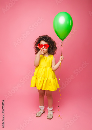 Happy birthday celebration with flying balloons of a charming cute little girl in a yellow dress isolated on a pink background.