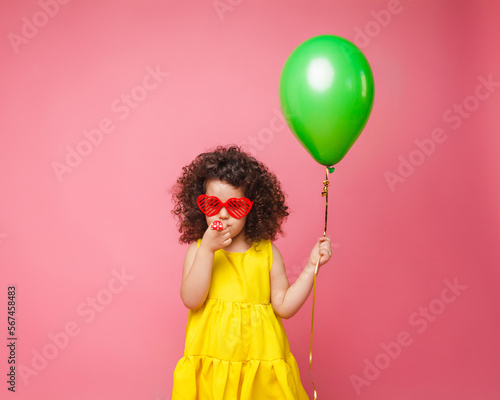 Happy birthday celebration with flying balloons of a charming cute little girl in a yellow dress isolated on a pink background.