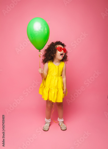 Happy birthday celebration with flying balloons of a charming cute little girl in a yellow dress isolated on a pink background. © inna717