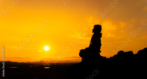 Person meditating on top of a mountain with the sun in front of him at sunset. Warm orange atmosphere. Silhouette of a man sitting on a rock. Copy space to the left.