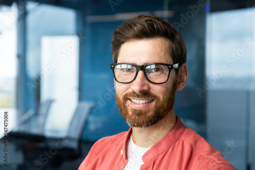 Close-up portrait of young businessman in red shirt, mature man in glasses smiling and looking at camera inside office near window, successful investor happy with achievements and good investment.