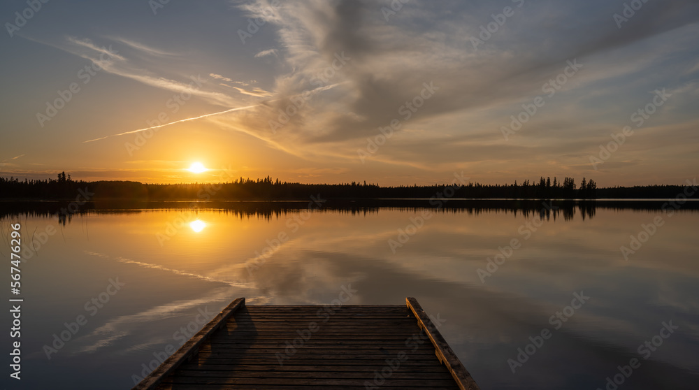 Colorful sunset above and reflecting in a calm lake with a dock in the foreground and evergreen trees along the lakeshore.  A plane vapor trail cuts through the sky.
