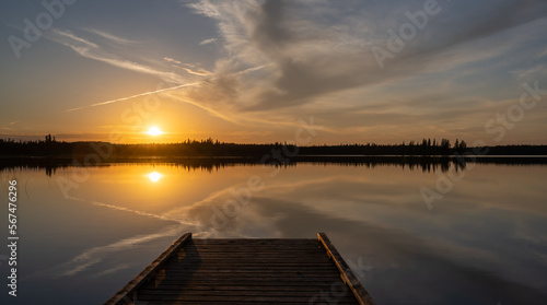 Colorful sunset above and reflecting in a calm lake with a dock in the foreground and evergreen trees along the lakeshore. A plane vapor trail cuts through the sky. 