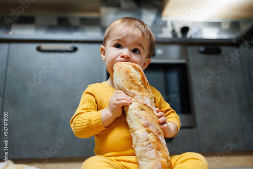 A cute one-year-old boy is sitting in the kitchen and eating a long bread or baguette in the kitchen. The first eating of bread by a child. Bread is good for children photo