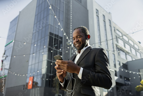 Photo from below. African American businessman in a suit standing outside an office center wearing headphones, using the phone. Listens, dials, calls, talks, smiles.