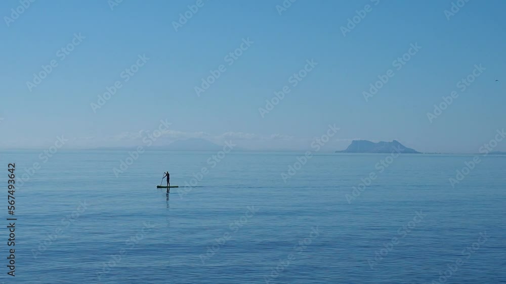 Summer scene in Andalusia, Spain, of the Mediterranean Sea with the Strait of Gibraltar in the background and unrecognizable vacationer practicing paddle surfing Summer scene in Andalusia, Spain, of the Mediterranean Sea with the Strait of Gibraltar in the background and unrecognizable vacationer practicing paddle surfing