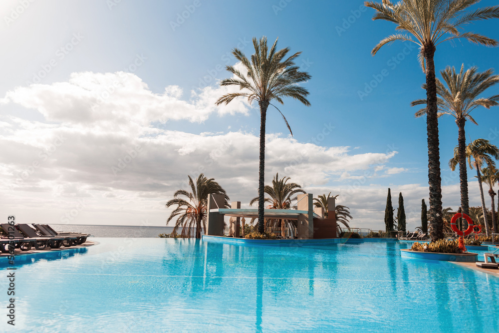 beautiful exotic view of a hotel with a pool, palm trees, sun beds and the ocean with a cloudy sky in Madeira island. Summer holidays in Funchal