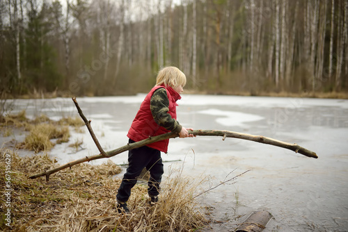Little boy playing large branch on shore of forest lake on early spring day. Surface of lake is still under ice. Child tries to break ice with stick. Outdoor activity for kids