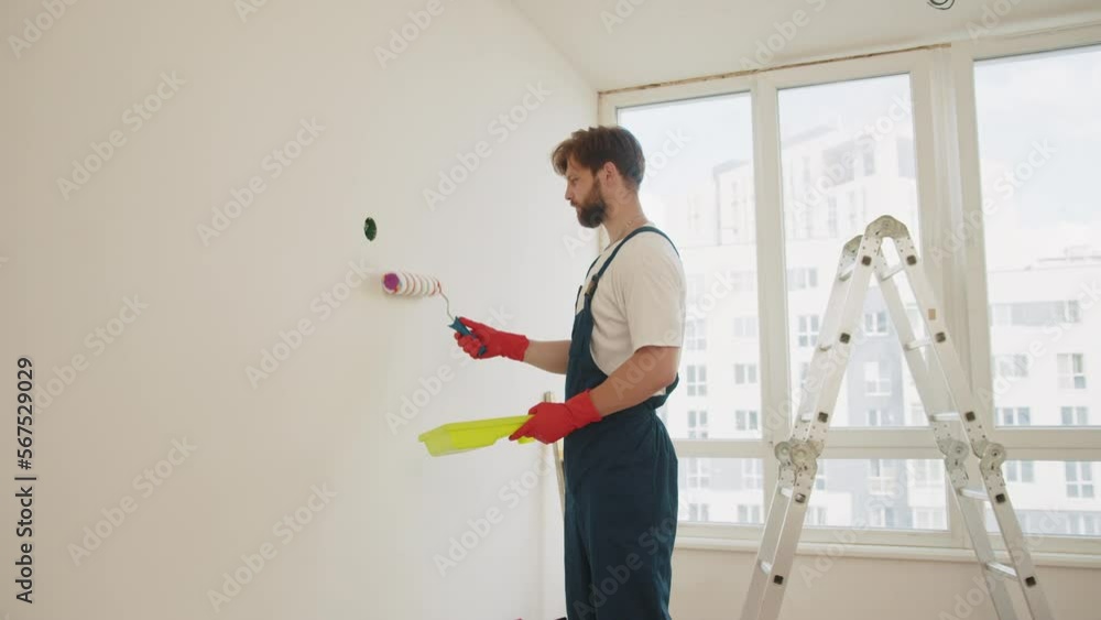 Close up of male in gloves holding painting roller. Painter man painting the wall in home, with paint roller and white color paint. Room renovations at house. Young worker painting wall in room. Close up of male in gloves holding painting roller. Painter man painting the wall in home, with paint roller and white color paint. Room renovations at house. Young worker painting wall in room.