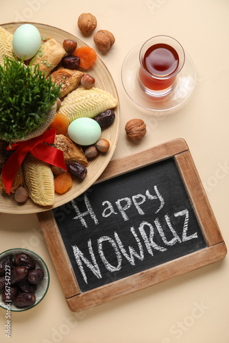 Chalkboard with text HAPPY NOWRUZ, treats, glass of tea and grass on beige background