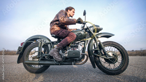 Side view of a stylish cool young man leather jacket driving his vintage motorcycle on an asphalt road at late autumn cloudy evening. © Nik Viatkin