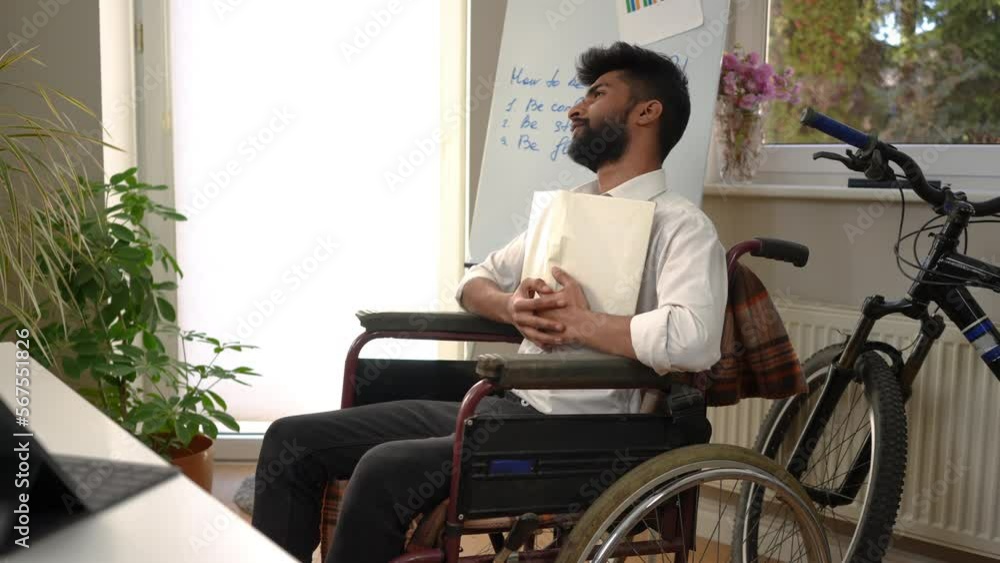 Handsome Middle Eastern intelligent young man reading hugging book looking away smiling sitting in wheelchair indoors. Side view portrait of positive satisfied freelancer enjoying hobby in home office Handsome Middle Eastern intelligent young man reading hugging book looking away smiling sitting in wheelchair indoors. Side view portrait of positive satisfied freelancer enjoying hobby in home office