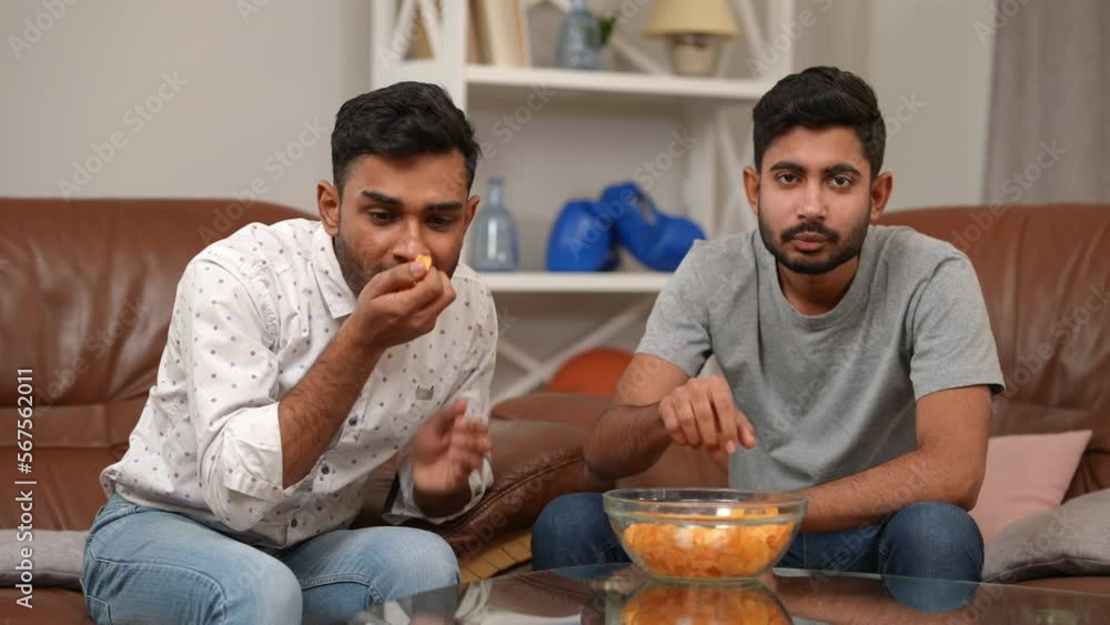 Two absorbed focused young Middle Eastern men eating chips looking at camera sitting on couch indoors. Concentrated millennial friends talking watching TV meeting on weekend Two absorbed focused young Middle Eastern men eating chips looking at camera sitting on couch indoors. Concentrated millennial friends talking watching TV meeting on weekend