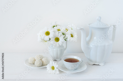 Fototapeta Naklejka Na Ścianę i Meble -  Tea and flowers. A bouquet of white daisies or chamomile iflowers in a vase on the table, a cup of tea, sweets and a teapot on a white background