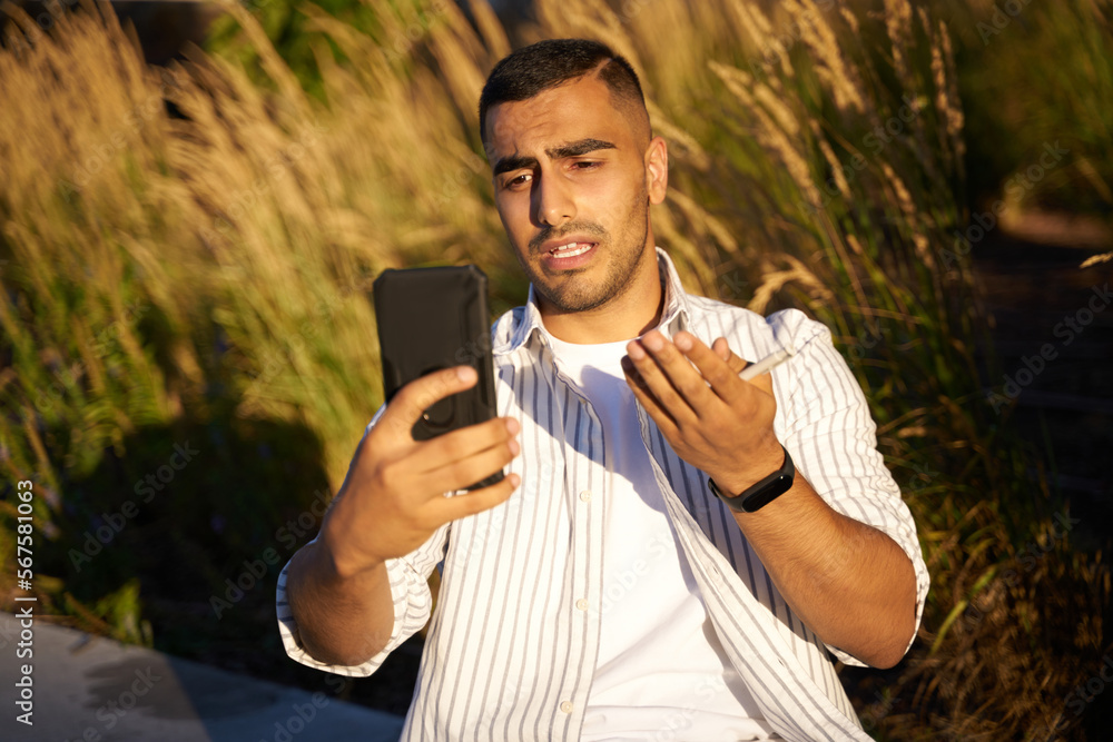 Middle eastern man holding mobile phone communication online, having video call sitting in park Middle eastern man holding mobile phone communication online, having video call sitting in park
