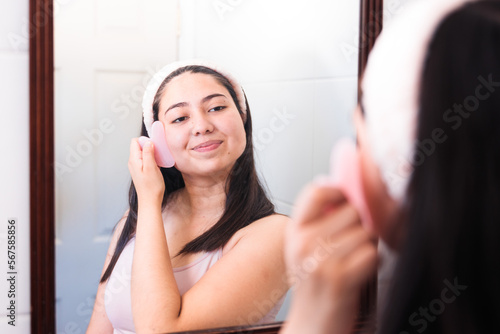 Smiling young woman in the mirror using gua sha quartz scraper stone to massage her cheek. Skin care