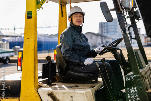 Middle-aged Asian male worker with forklift on factory grounds.