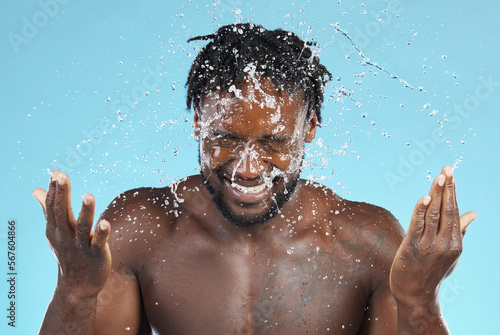 Water splash, cleaning and hygiene with a model black man in studio on a blue background for hydration. Bathroom, skincare and wellness with a young male wahsing his face for natural skin treatment photo