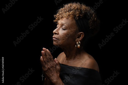 Beautiful elderly 70+ year old Christian African American black woman religiously and fervently praying to God with eyes closed and hands together holding a cross