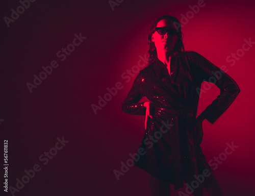 Young beautiful confident brunette female in trendy evening shiny green reflecting dress. Sexy carefree woman isolated on red neon light in studio. Fashionable model in sunglasses. Fantasy style