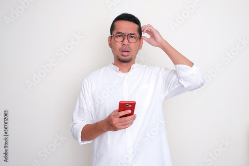 Adult Asian man showing confused expression while holding mobile phone photo