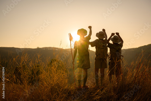 three male students hiking with backpack Standing with hands raised in joy on the mountain Boy Scouts rejoice at rock climbing success in the evening at sunset.