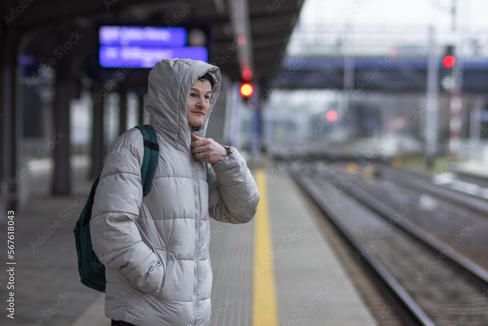 Fototapeta premium Thoughtful young man in casual gray jacket and hat with backpack standing and waiting for train at railway station.