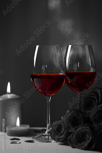 Glasses of wine, burning candles and rose flowers for romantic dinner on grey table against blurred lights. Black and white effect with red accent