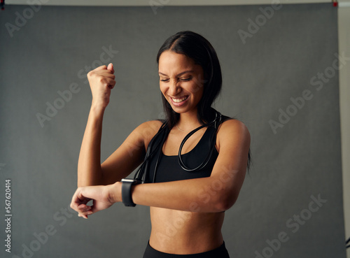 Happy young biracial woman in sports bra smiling while checking the time on fitness tracker in studio