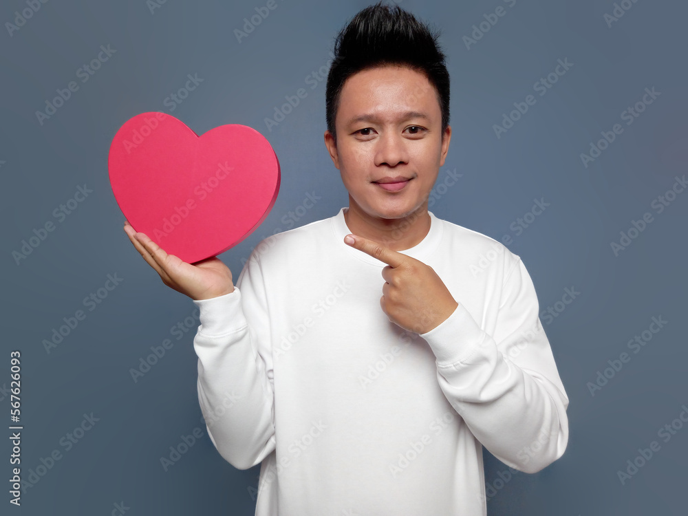 Handsome man wearing white long sleeve sweater holding red heart shape and pointing. Handsome man wearing white long sleeve sweater holding red heart shape and pointing.
