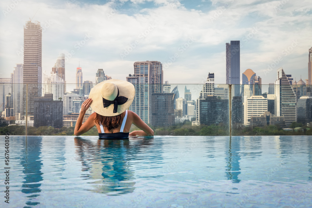 Fototapeta premium A woman with hat enjoys the elevated view over the urban skyline of Bangkok, Thailand, from a swimming pool