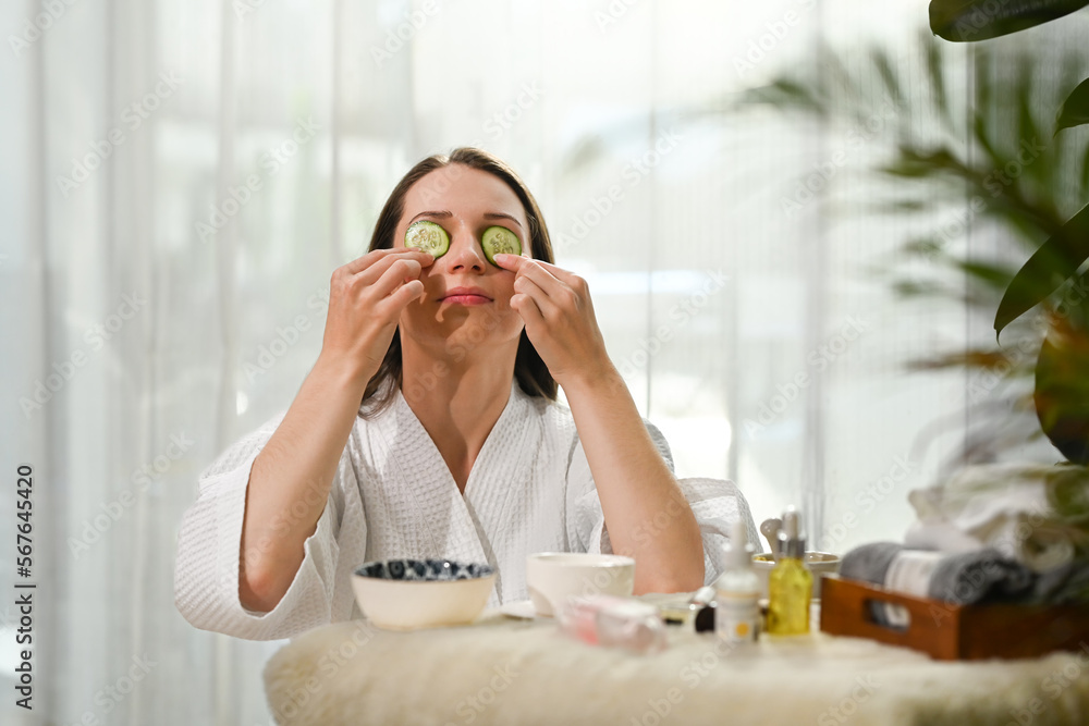 Young woman in bathrobe with cucumber slices on eyes, taking spa skincare procedures. Beauty treatment and detox concept Young woman in bathrobe with cucumber slices on eyes, taking spa skincare procedures. Beauty treatment and detox concept