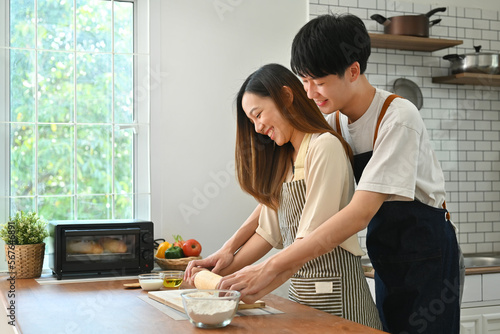 Attractive man and woman in casual outfits kneading dough with a rolling pin on wooden table in kitchen interior