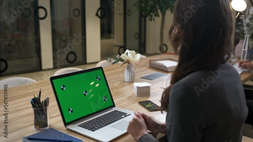 A woman at a office desk works at a laptop. The concept of a successful business woman, a leader, feminism. Online negotiations with the interlocutor online using a messenger. Green screen mockup.