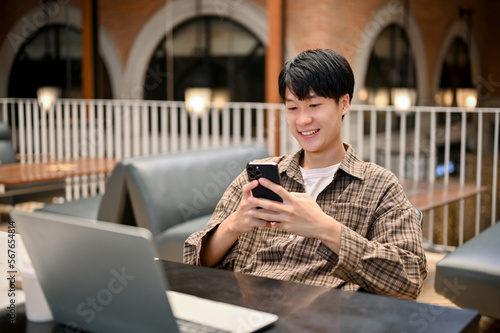 Relaxed Asian male freelancer or college student using his smartphone, remote working at cafe